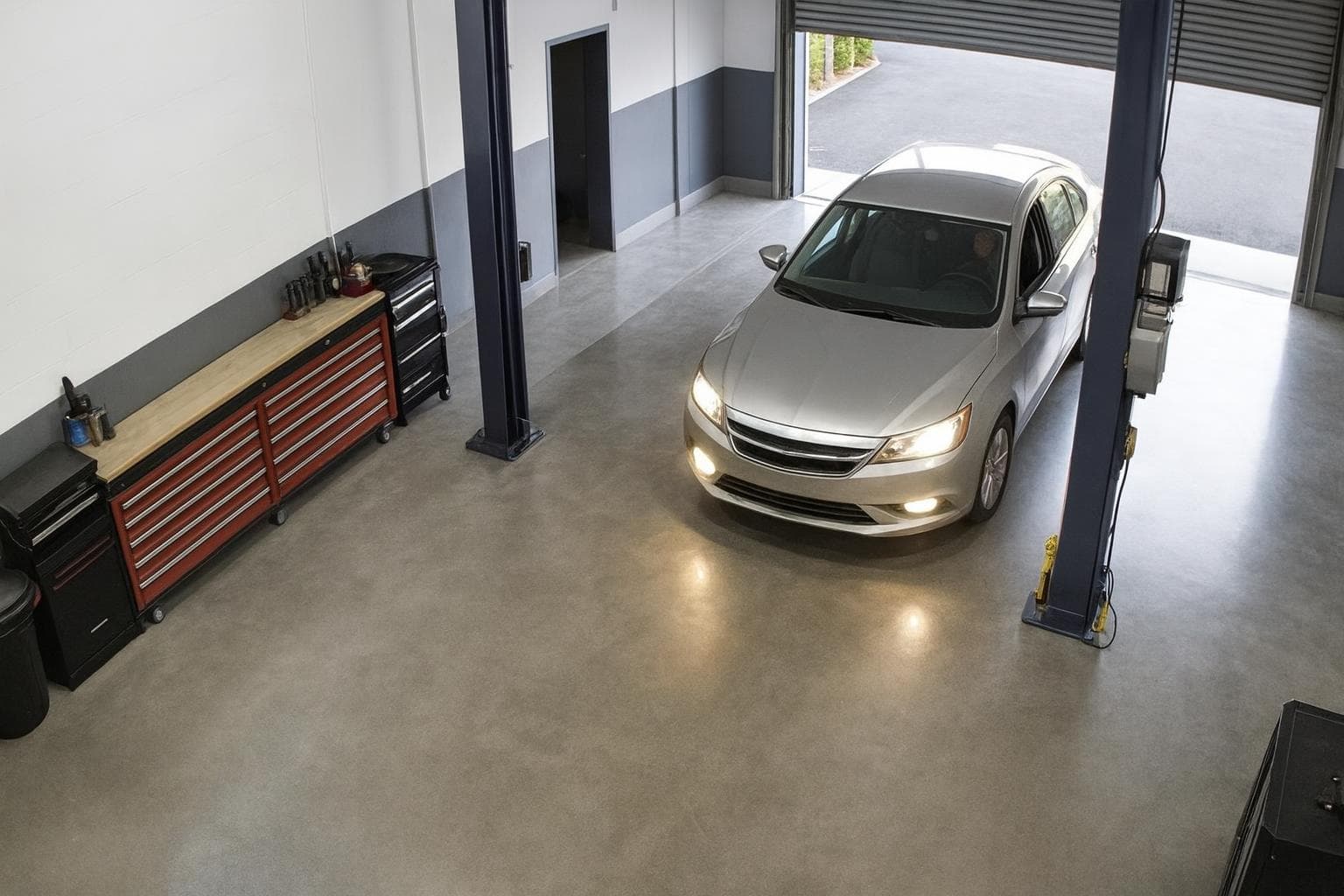 Service bay camera view of a silver sedan driving in through the roller door, headlights on